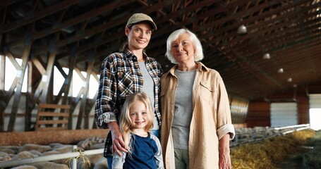 Portrait of Caucasian cheerful mother, old grandmother and cute small pretty girl in stable with sheep smiling to camera. senior woman with daughter and granddaughter standing in barn with livestock.