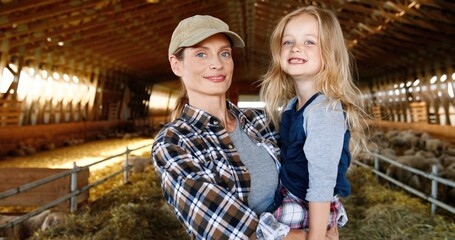 Caucasian beautiful woman holding small cute pretty daughter on hands and smiling to camera in stable with sheep flock. Indoor. Little girl with lovely mother. Farming at countryside. Motherhood love.
