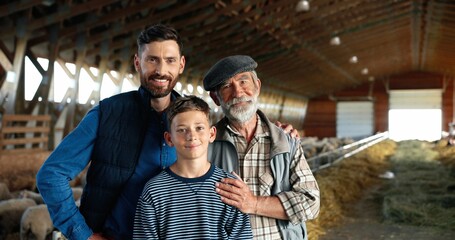 Obraz na płótnie Canvas Three male generations in green field at cattle farm. Caucasian grandfather, father and son smiling to camera and standing at stable with sheep flock. Shepherds. Old man farmer with son and grandson.