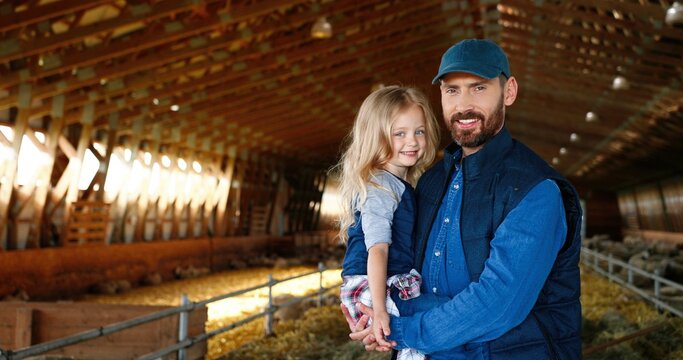 Portrait of Caucasian handsome father holding on hands cute little daughter and smiling to camera at sheep stable in farm. Farming lifestyle. Countryside concept. Smiled man and small girl indoors.
