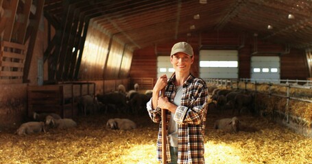 Portrait of beautiful Caucasian young woman in hat looking at camera with smile and leaning on stick of pitchfork. Pretty female shepherd smiling in stable. Dolly shot. © VAKSMANV