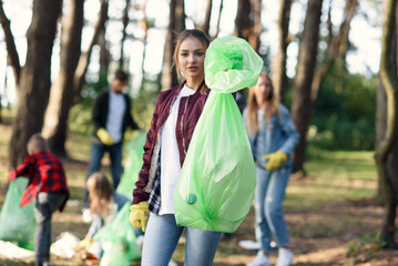 Pretty young woman shows a full rubbish pack in background of his friends volunteers picking up...