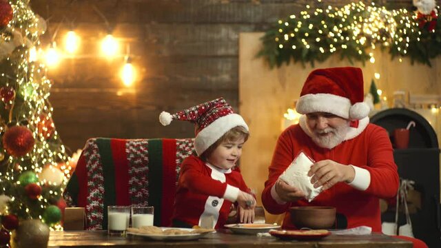 Kid and Santa Claus enjoying in served gingerbread cake. Santa Grandfather and grandson having fun near Christmas tree and celebrate Christmas at home. Funny cute cild make Christmas cookies.