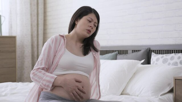 portrait woman in the third trimester is stroking her belly with a painful look while having severe contraction pain in the morning on bed at home.