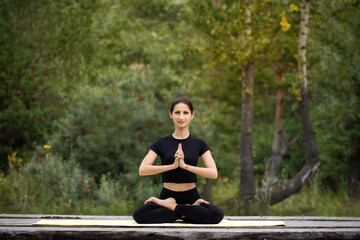 a woman is doing yoga and smiling cheerfully in the forest on a wooden deck sitting in the Lotus position. She is sitting in the Lotus position