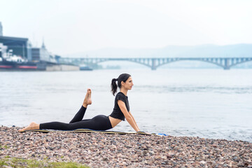 Fototapeta na wymiar a woman does yoga next to a river and against the background of a bridge and city buildings