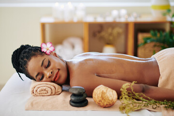 Fototapeta na wymiar Beautiful young Black woman with flower in her hair resting on bed in spa salon after relaxing back massage with oils