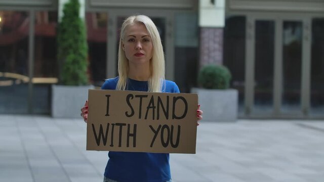 Young woman blonde stands with a cardboard poster I STAND WITH YOU in a public place outdoor. Single protest. No racism, tolerance and political correctness in society. Slow motion. Close up.