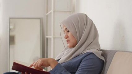Beautiful Asian muslim woman reading book while sitting on sofa, girl enjoys her time by doing leisure activity, happy cheerful expression