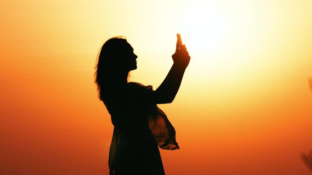silhouette woman holding smartphone using camera taking photos at sunrise warm sky with rising sun