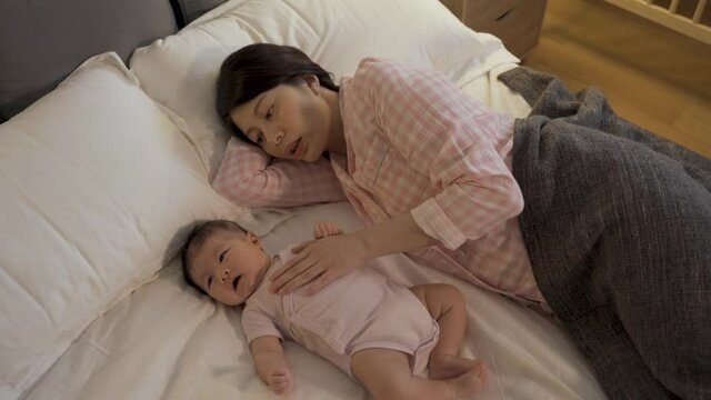high angle shot japanese mom lying on her side in the bedroom with hand under head is patting on baby’s chest to help her calm down and fall asleep.