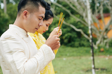Asian couple in traditional costumes praying with incense sticks in hands