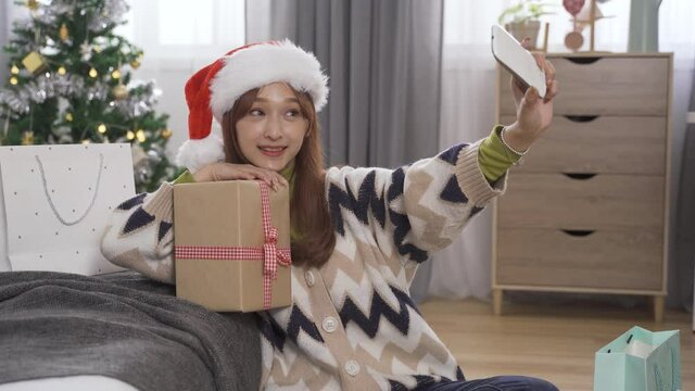 asian happy girl with Christmas cap sitting by bedside at home is texting and sharing photo after taking selfie by leaning her head on the gift.
