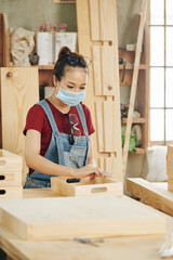 Pretty young female carpenter in protective mask sanding surface of wooden crate