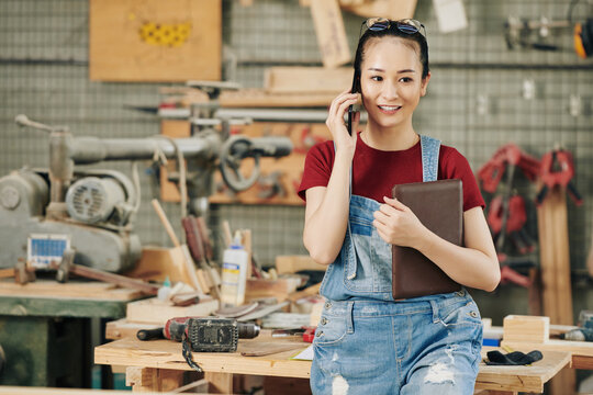 Cheerful young female carpenter in denim overall holding tablet computer and calling on phone