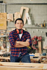Portrait of confident mature carpenter with earmuffs standing with arms folded in his workshop