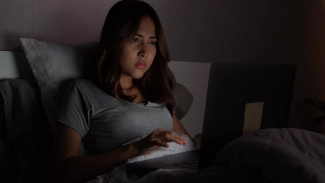 Young Asian woman lying in the bed and working with a laptop late at night, suffering and exhausted.