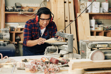 Skilled mature Asian craftsman in earmuffs cutting wooden plank with saw in a workshop