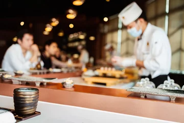 Selbstklebende Fototapeten Japanese Omakase Restaurant counter focuses on Japanese ceramic tea cup with Blur chef cooking at the kitchen counter and directly serve to customers in the background. © artitwpd