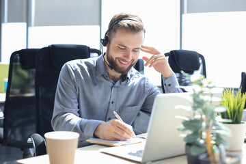 Smiling male call-center operator with headphones sitting at modern office, consulting online...