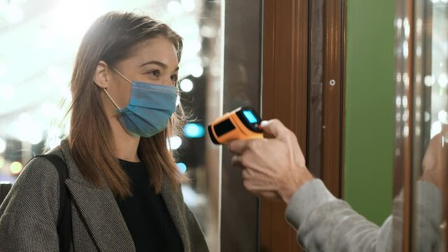 Taking body temperature before entering a restaurant. Screening of people for Covid-19 coronavirus symptoms. Young woman in medical protective mask enters the store