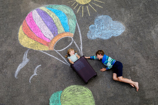 Happy little toddler girl and kid boy flying in hot air balloon painted with colorful chalks in rainbow colors on ground or asphalt in summer. Two children, siblings having fun. Creative leisure