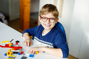 Naklejka na ściany i meble Little blond child with eye glasses playing with lots of colorful plastic blocks. Adorable school kid boy having fun with building and creating robot. Creative leisure modern technic and robotic.