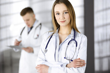 Fototapeta premium Professional beautiful woman-doctor with a stethoscope is standing with crossed arms in a clinic. Young doctors at work in a hospital. Medicine and healthcare concept