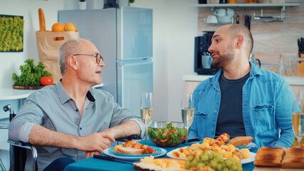 Middle aged man and older senior in wheelchair having fun during family dinner. Happy men smiling, drinking and eating during a gourmet meal, enjoying time sitting around the table in the kitchen