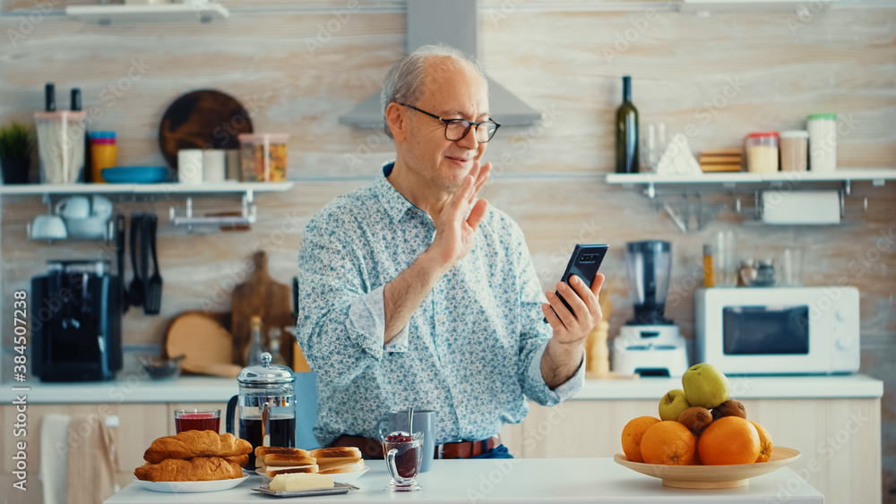 Poster senior man shacking hand during a video call in kitchen using smartphone. elderly person using inter