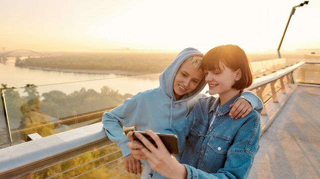 Lesbian couple standing on the bridge, smiling while taking a selfie picture, watching the sunrise together