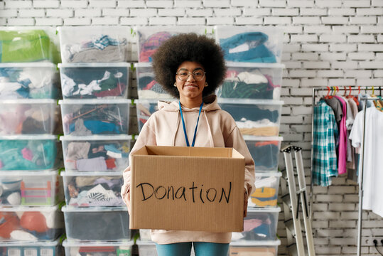 African american woman holding donation box and smiling at camera, posing in front of boxes full of clothes, Young volunteer working for a charity