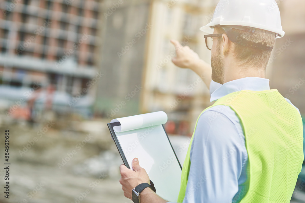 Wall mural building industry. rear view of a young civil engineer or construction supervisor wearing helmet poi