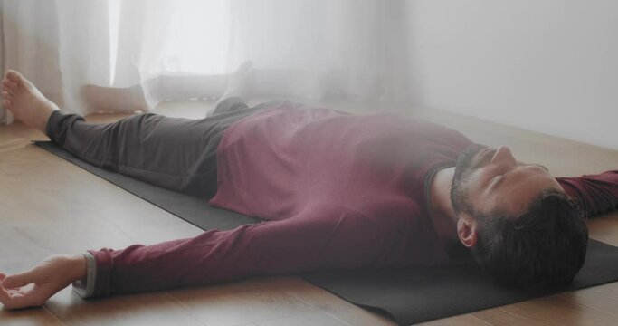 Low angle view of man relaxing in shavasana asana indoors natural morning light. Bearded Caucasian instructor lying on yoga mat after practice slow motion. relaxation meditation stress overcome