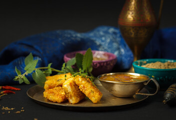 Cheese in mint batter with chutney sauce, an Indian dish. On a copper plate with fresh mint, bowls of spices in the background. Low key, with space