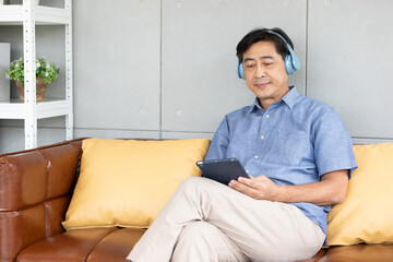 senior asian man wearing headphones and enjoy favorite music from tablet on sofa