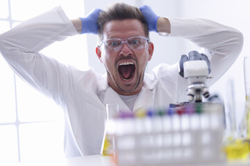 Man in goggles and rubber gloves pulls out hair on his head in chemical laboratory portrait. Mad...