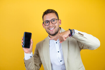 Young handsome businessman wearing suit over isolated yellow background smiling and pointing the phone