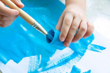 A child's hand holds and dips the brush in the paint. Smears of blue paint on white paper.
