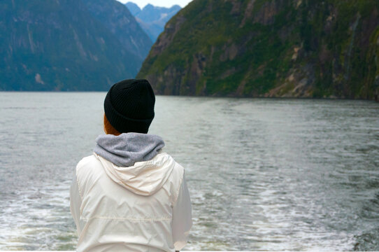 View From The Back Of Unrecognisable Woman Traveller In White Coat Looking Over Beautiful Lake And Mountains From Tourist Boat At Milford Sound In New Zealand