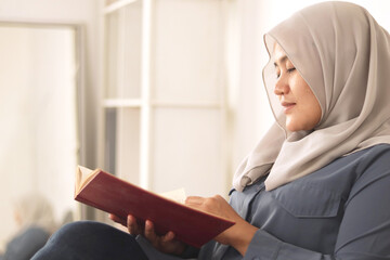 Beautiful Asian muslim woman reading book while sitting on sofa, girl enjoys her time by doing leisure activity, happy expression