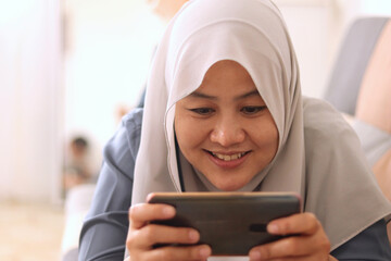 Beautiful Asian muslim woman using phone while relaxing on sofa, playing games or watching...