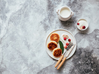 Cottage cheese pancakes on white plate with sour cream and berries on grey background. Homemade traditional Ukrainian and Russian syrniki. Healthy cheesecakes for breakfast. Top view with copy space.