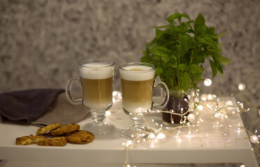 Hot chocolate cappuccino latte coffee with froth in a glass cup mug with cookies. White tray with hot drinks, garland, bokeh.