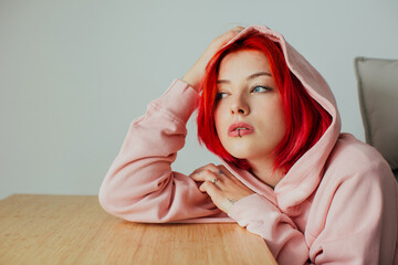 Portrait of a cool teen girl with bright red hair, lip piercing and hoodie holding head on desk...