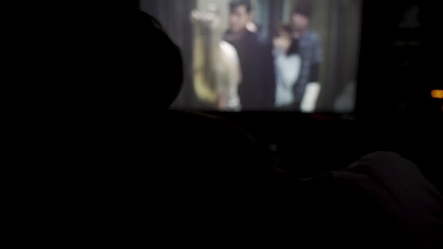 Rear view of a romantic couple in cinema with the blurred movie on the background. Media. Silhouettes of loving couple watching movie, woman putting her head on male shoulder.