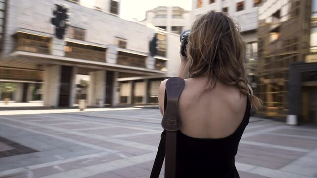 Beautiful young photographer woman takes photos outdoors of a modern business district. Action. Camera moving around a beautiful slim woman in black dress with a professional camera in her hands.