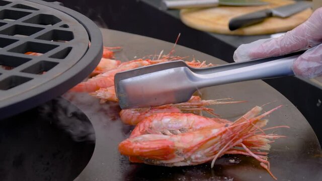 Seafood, barbecue, outdoor cooking, gastronomy, cookery, street food concept. Slow motion: chef grilling fresh red king prawns on round black brazier at summer local food market - close up view