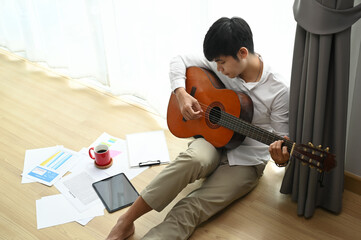 Young man sitting on floor and holding acoustic instrumental guitar playing new written song .