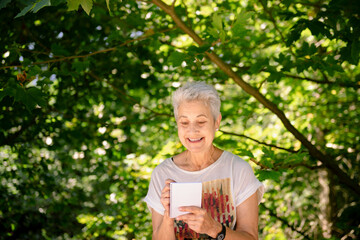 Senior woman hiker takes notes contemplating nature. she is smiling enjoying a sunny day among the forest trees.